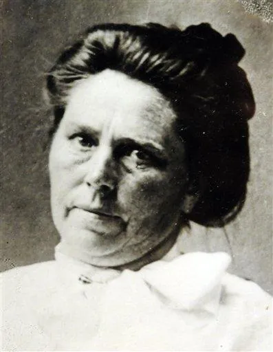 Black and white photo of a woman with tied back hair, wearing a high-collared white blouse, looking at the camera.